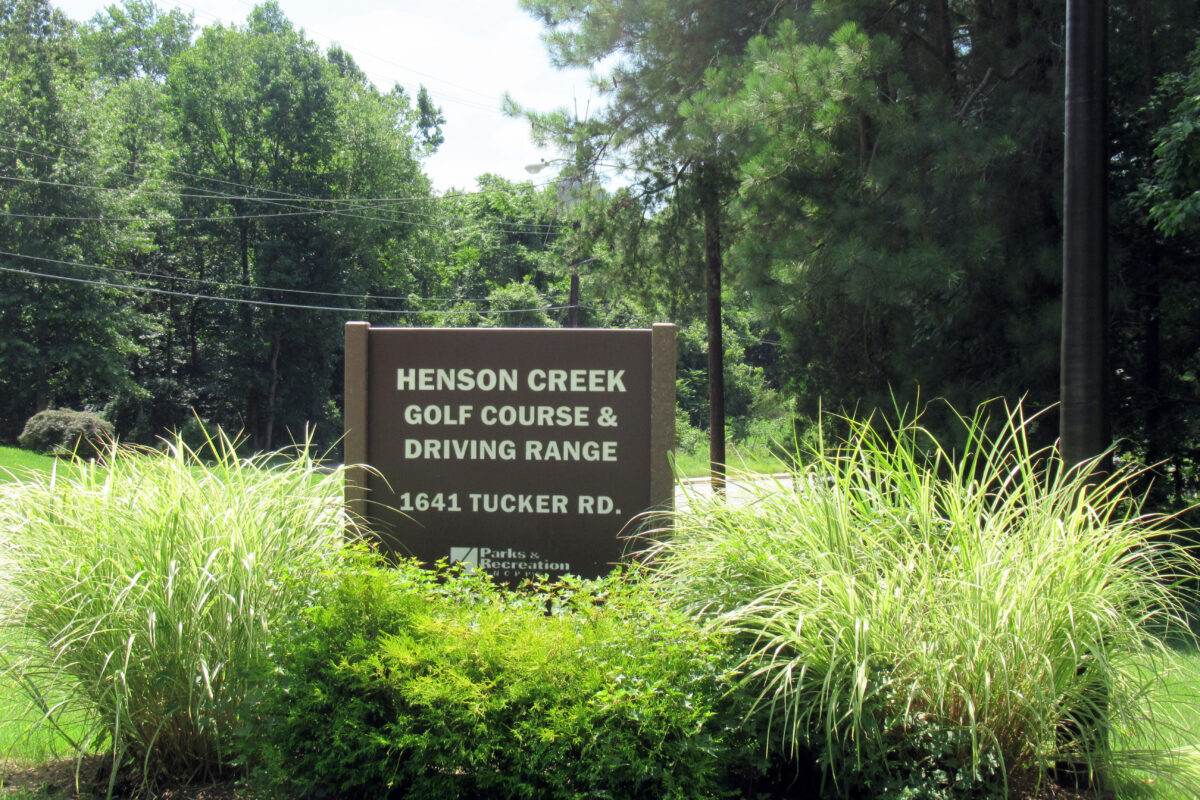 A brown sign mounted in the ground surrounded by tall ornamental grasses. The sign reads HENSON CREEK GOLF COURSE & DRIVING RANGE. A road and trees are visible in the distance.