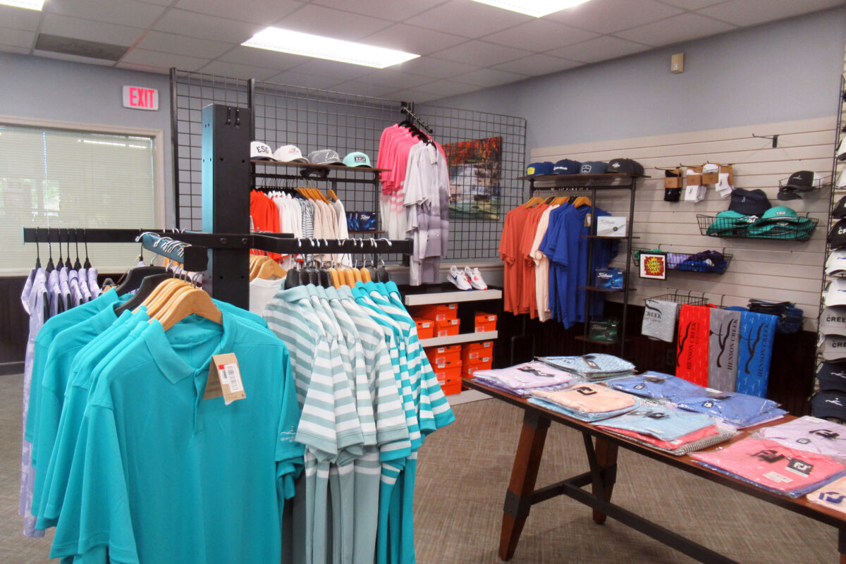 A small retail area containing several racks of colorful polo shirts, hats, and a table containing folded clothing items. Shelves near the floor are stacked with orange shoe boxes.