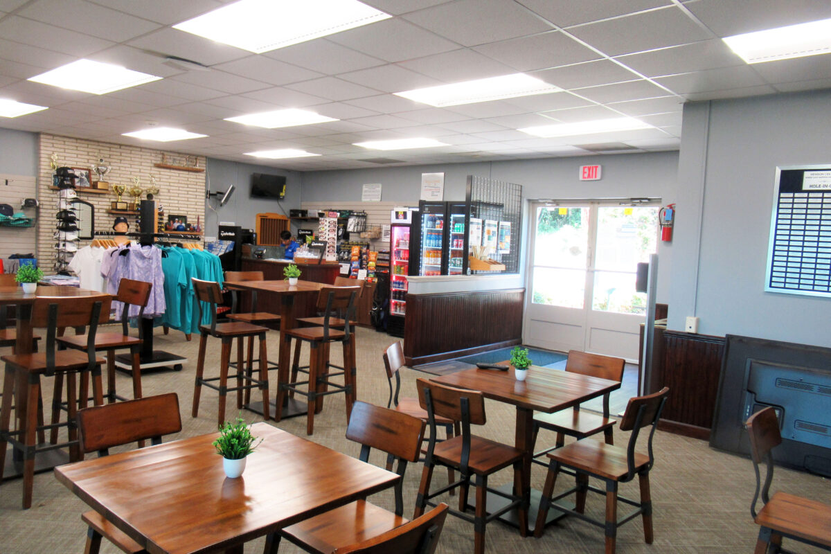 An interior space with a small retail area in the rear and several wooden cafe tables with chairs in the foreground. Glass double doors in the far wall lead outside.