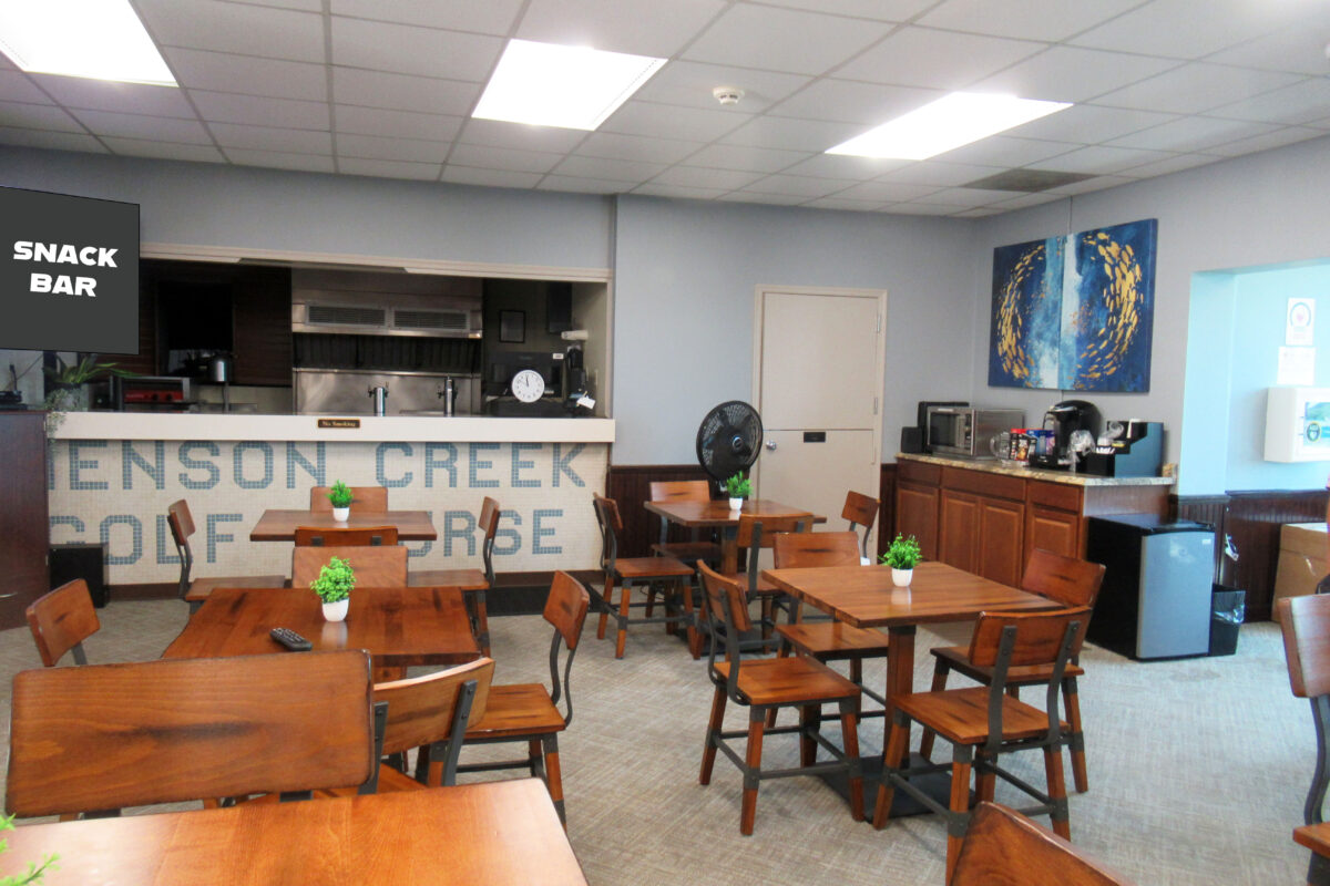 Half a dozen wooden cafe tables with chairs arranged in a room. There is a counter in the far wall with tilework that reads HENSON CREEK GOLF COURSE. The words SNACK BAR are superimposed over the photo.