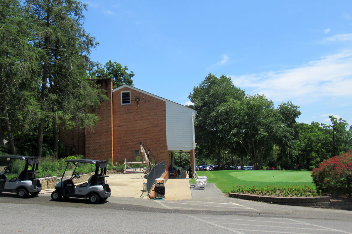 Two golf carts are parked near the edge of a concrete patio that extends out from a two-story brick building. In front of the building is a leveled putting green. A row of low chairs along the building face the green.