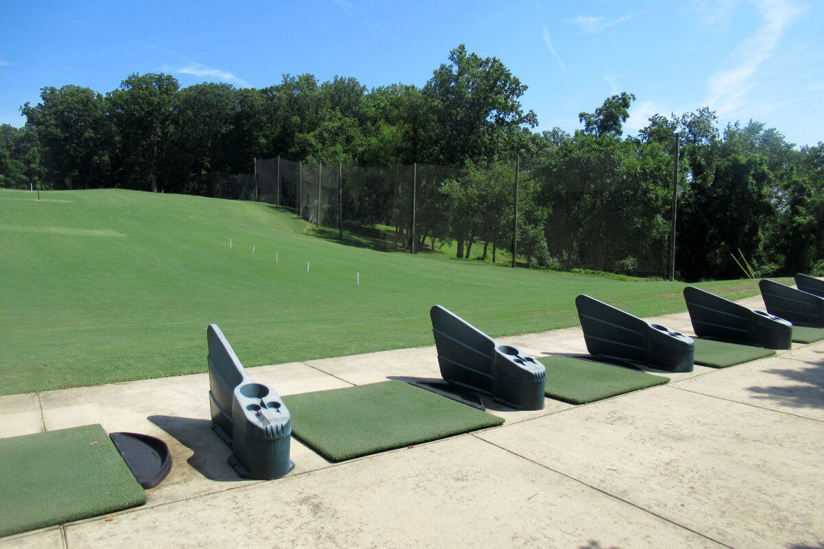 The teeing area of a driving range, showing about six teeing boxes separated by small, low walls. They face a large expanse of green grass that gradually slopes upward. This area is enclosed on the far side by a tall fence.