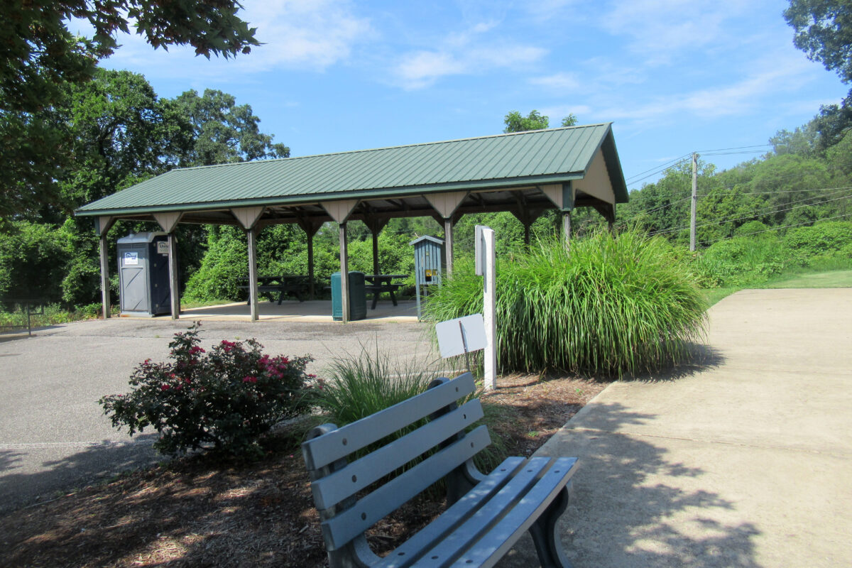 A picnic shelter with a green, peaked roof covering several wooden picnic tables on a concrete pad. An asphalt parking area adjoins the structure, and a concrete paved path runs alongside it. There is a porta-john under the shelter at the far end.
