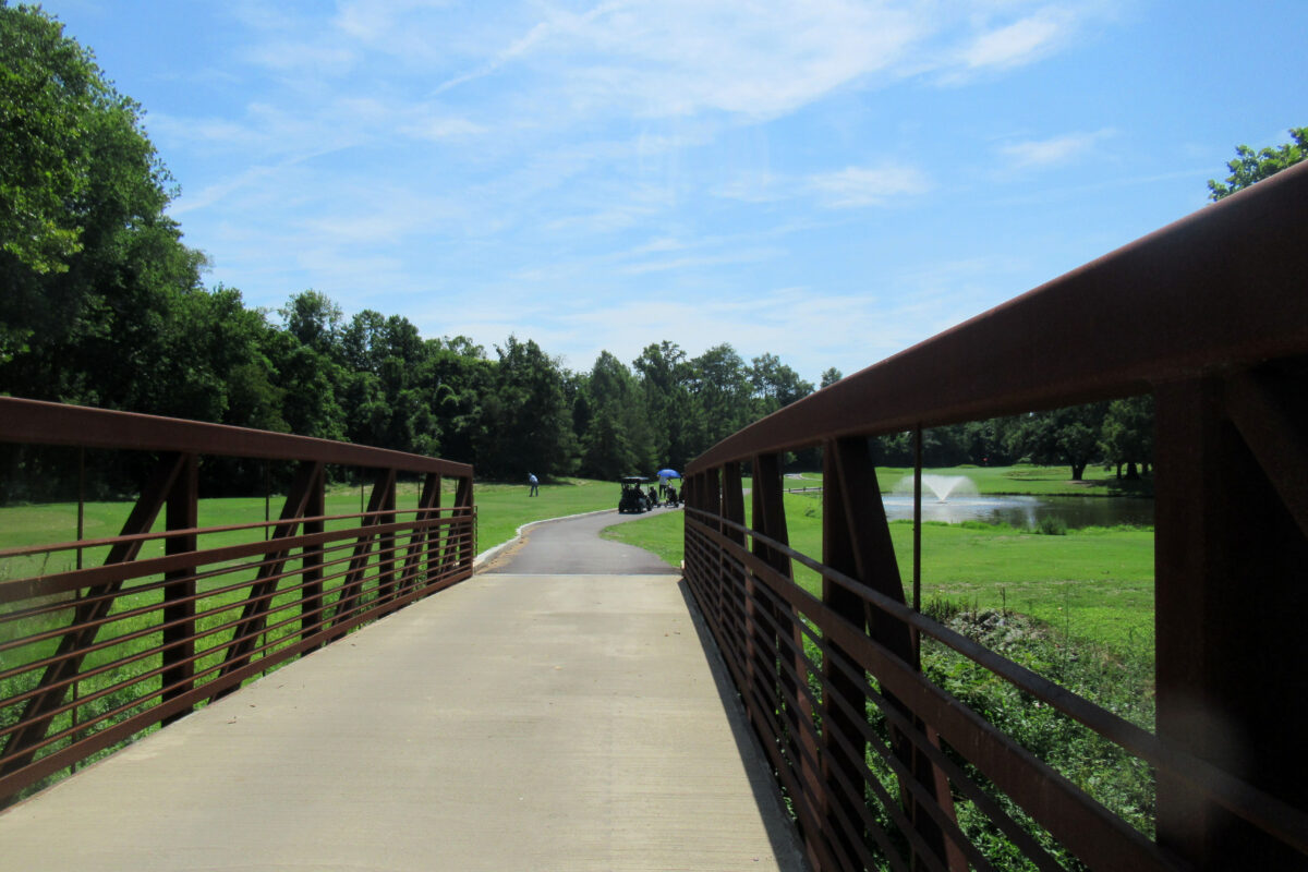 A concrete and metal trestle bridge leading to a paved path with several golf carts on it. The path runs through an open, grassy area and past a small pond with a fountain of water spraying up in the middle.