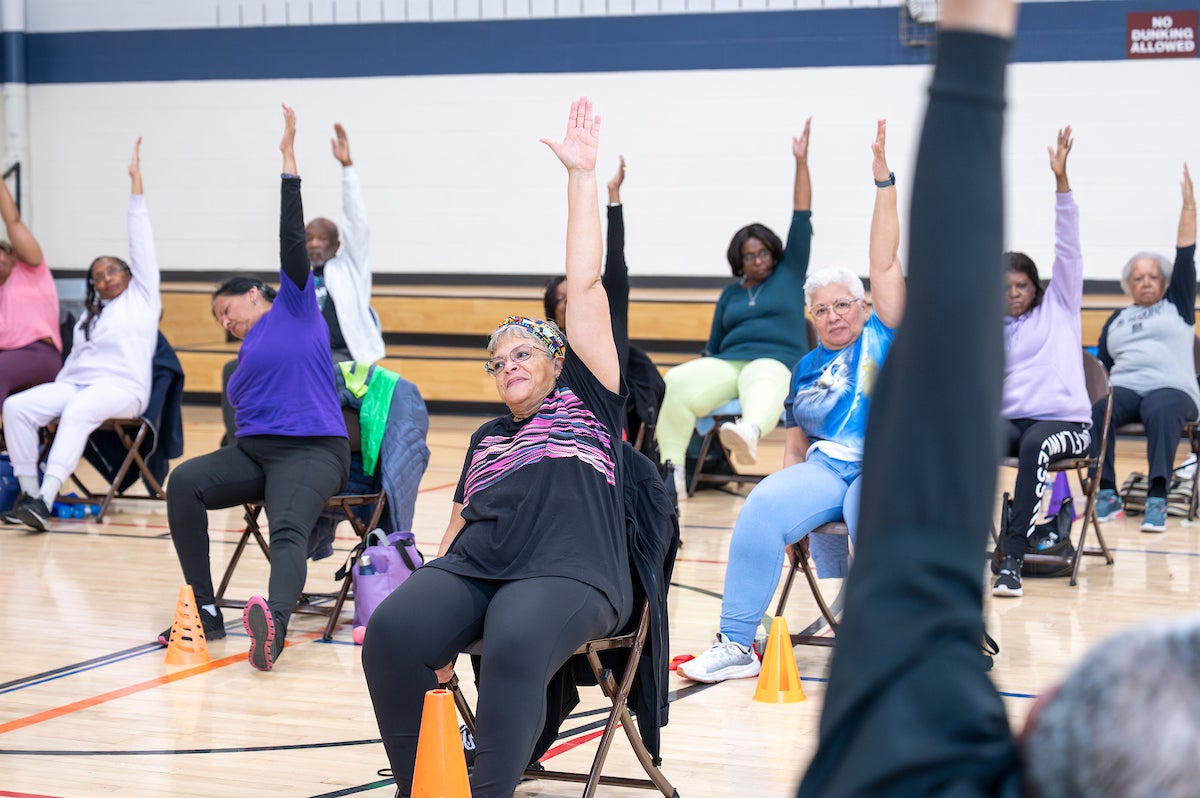In a gymnasium, about a dozen seniors sitting in folding chairs all reach their left arms upwards in a stretch. The instructor can just be seen in the corner of the image, at the front of the exercise class.