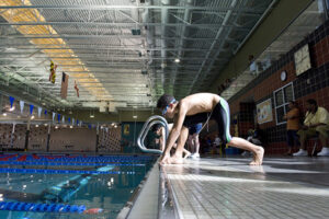 Side view of a three young men poised in position along the side of a large indoor pool, ready to dive into their swimming lanes.