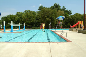 An outdoor pool on a sunny day with play areas including yellow and red waterslides. A lifeguard sits in a chair under a blue and white umbrella.