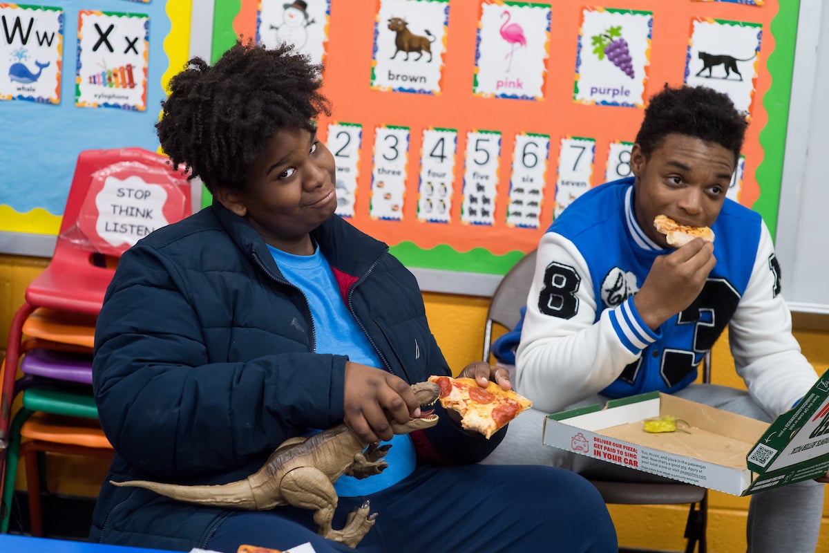 In a classroom with educational bulletin boards, two teens sit on folding chairs eating pizza. One looks comically at the camera as he feeds his slice of pizza to a plastic dinosaur toy.