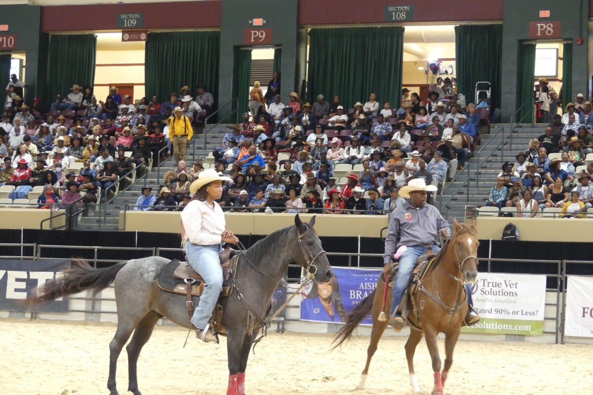 Two cowboys wearing hats ride horses across a sandy arena floor in front of spectating arena bleachers.