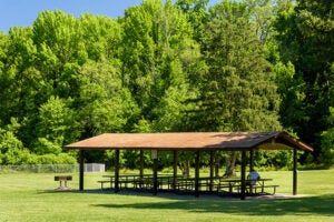 A picnic shelter in a grassy lawn with four picnic tables under it, on a concrete patio. There is a grill installed in the grass nearby. Past the far edge of the grass are tall trees.