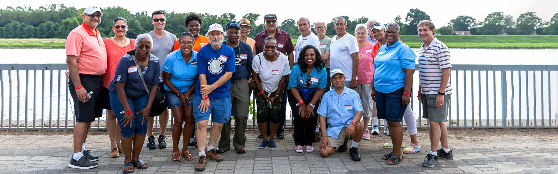 Twenty adults pose standing on a concrete patio in front of a metal railing. They all wear nametag stickers. Behind them, on the other side of the metal railing, is a large body of water.
