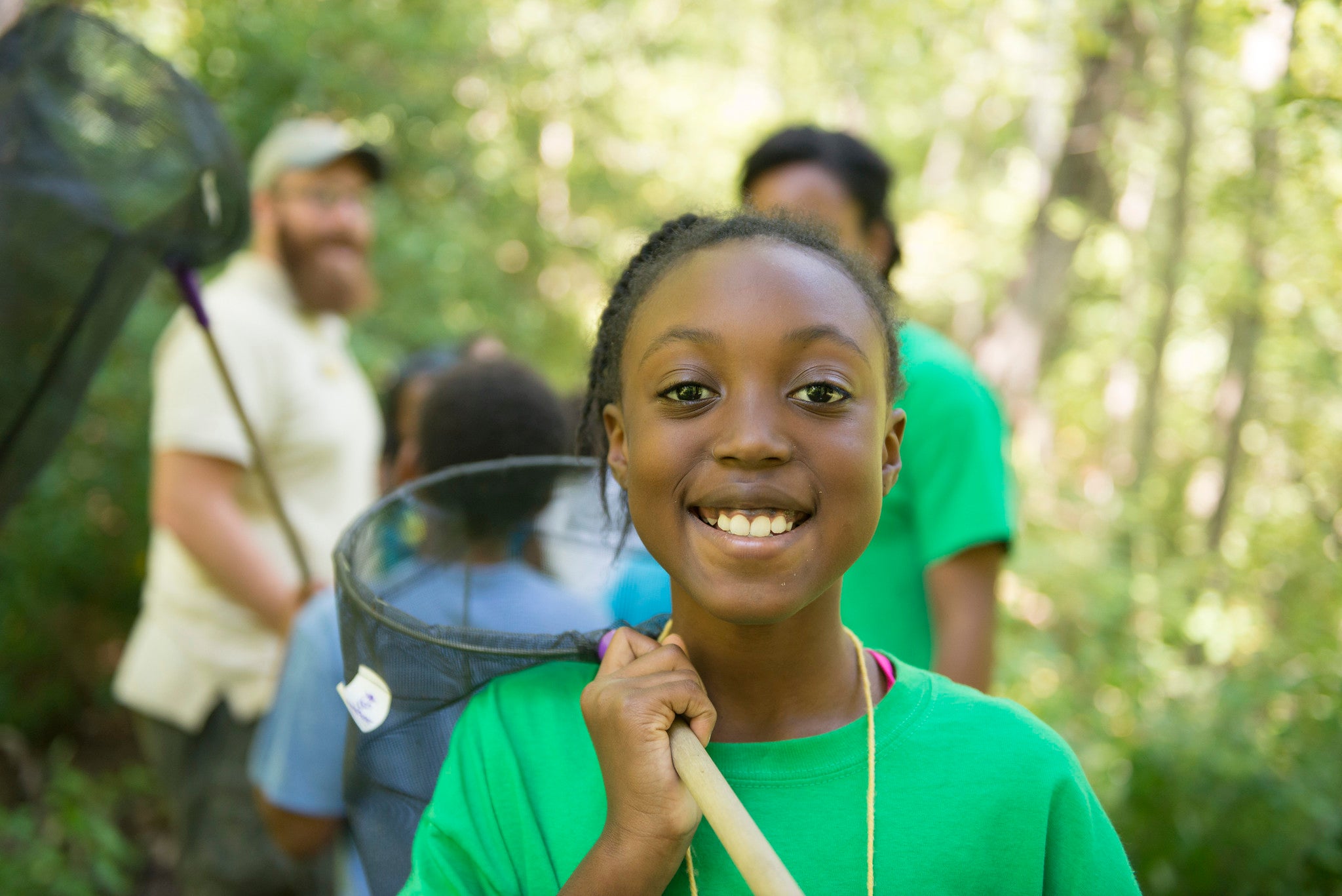 Close-up of a smiling girl in a green shirt holding a net on a long pole over her shoulder. Behind her is a wooded area.