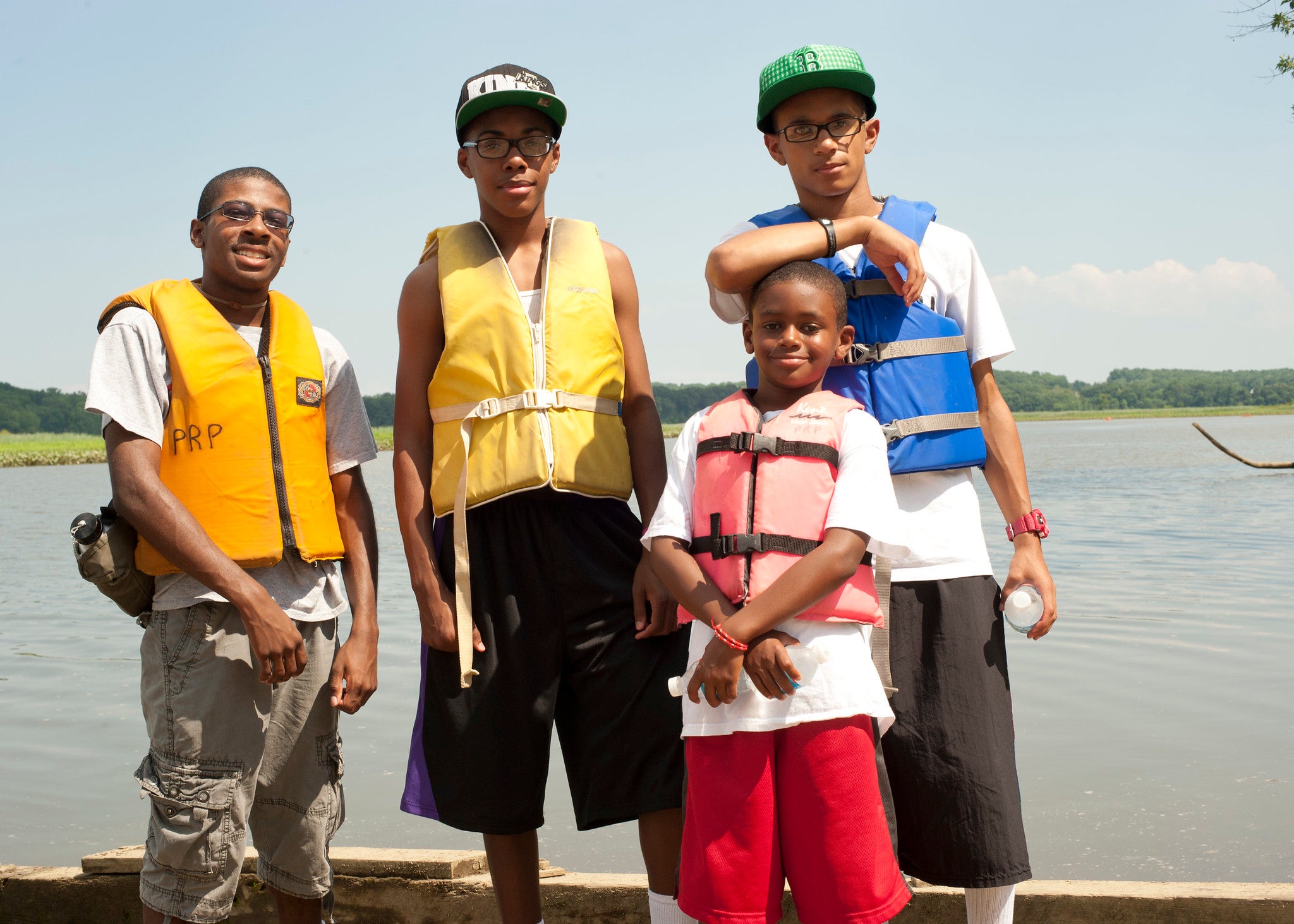 Four youths pose smiling in life vests on a dock next to a large body of water.