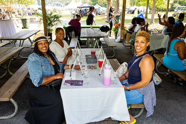 Three smiling women sit at a picnic table under a picnic shelter, each with a wine glass and a small easel in front of her. In the distance is a grassy field with many event tents, and a crowd of people.