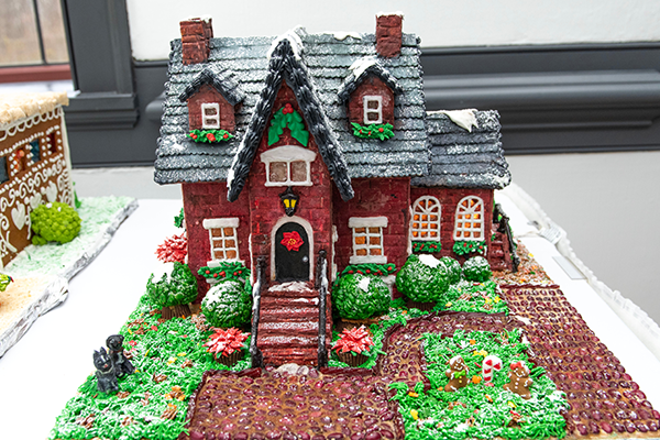 A red brick gingerbread house with icing-lined shingles, candy window panes, frosting grass, peanut butter cup bushes and a driveway and walkway made out of cobblestone coffee beans.