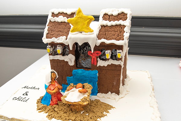 A gingerbread house with white frosting decorations, blue stairs leading to the door, and a giant yellow star on top of the roof. Below sits a mother kneeling over her baby lying in a manger.
