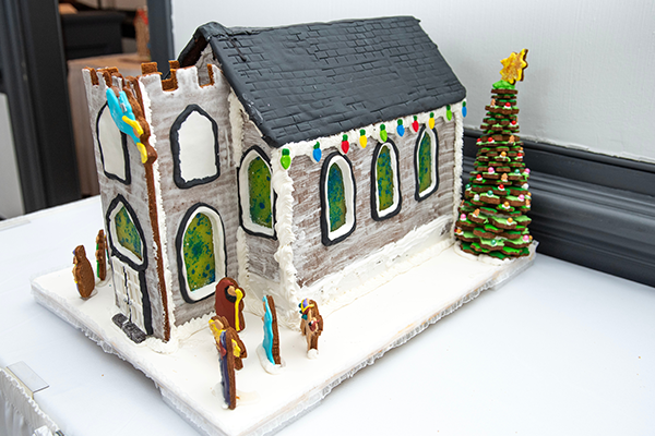 A gray gingerbread church with white icing mortar with a large front steeple and green and blue sugar stained glass windows. There is a nativity scene out front, and a gingerbread tree at the back corner of the building.