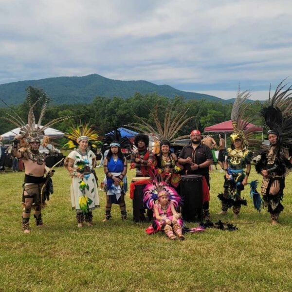 Nine people pose in a grassy field wearing elaborate costumes and large, feather headdresses. Two of them are standing behind large drums. In the background, several event tents are set up in a row.