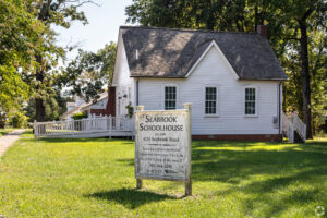 A white wooden sign reading Seabrook Schoolhouse sits in a green yard. Behind it is a small white schoolhouse with clapboard siding and a steep gray roof.
