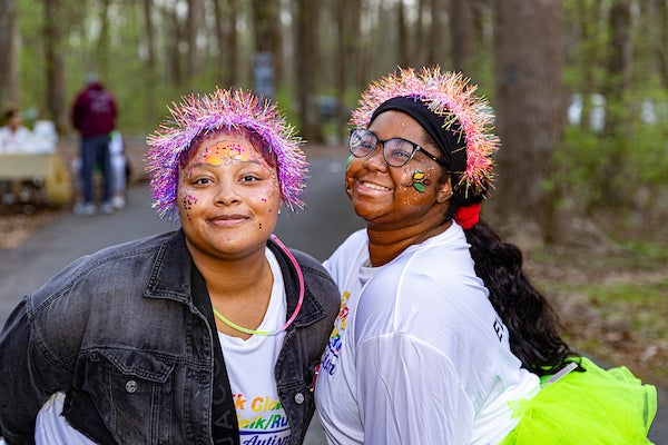 Two women with face paint and tinsel garland headbands smile on a paved road, wearing neon and glow sticks.