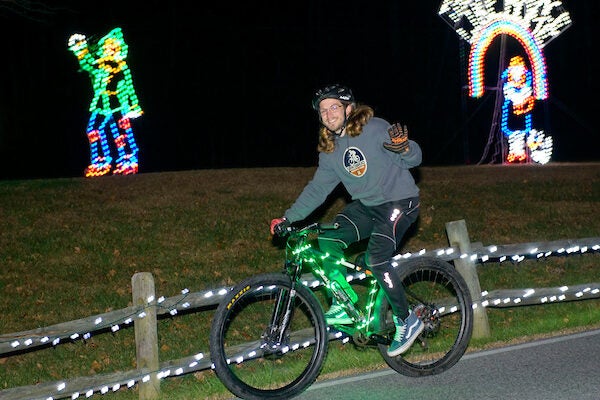 Person in grey sweater riding bike while waveing. They are passing a person made of holiday lights