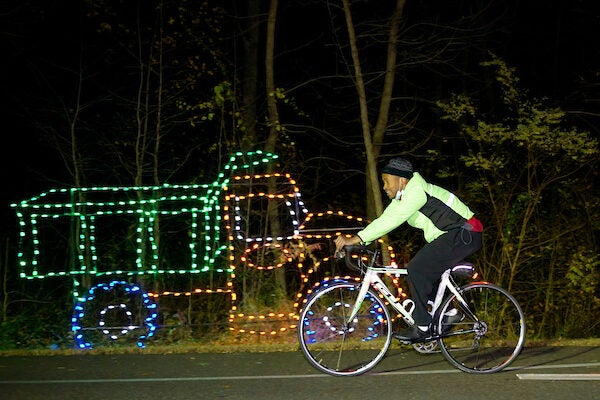 Person in green jacket riding bike pass truck made of Christmas lights
