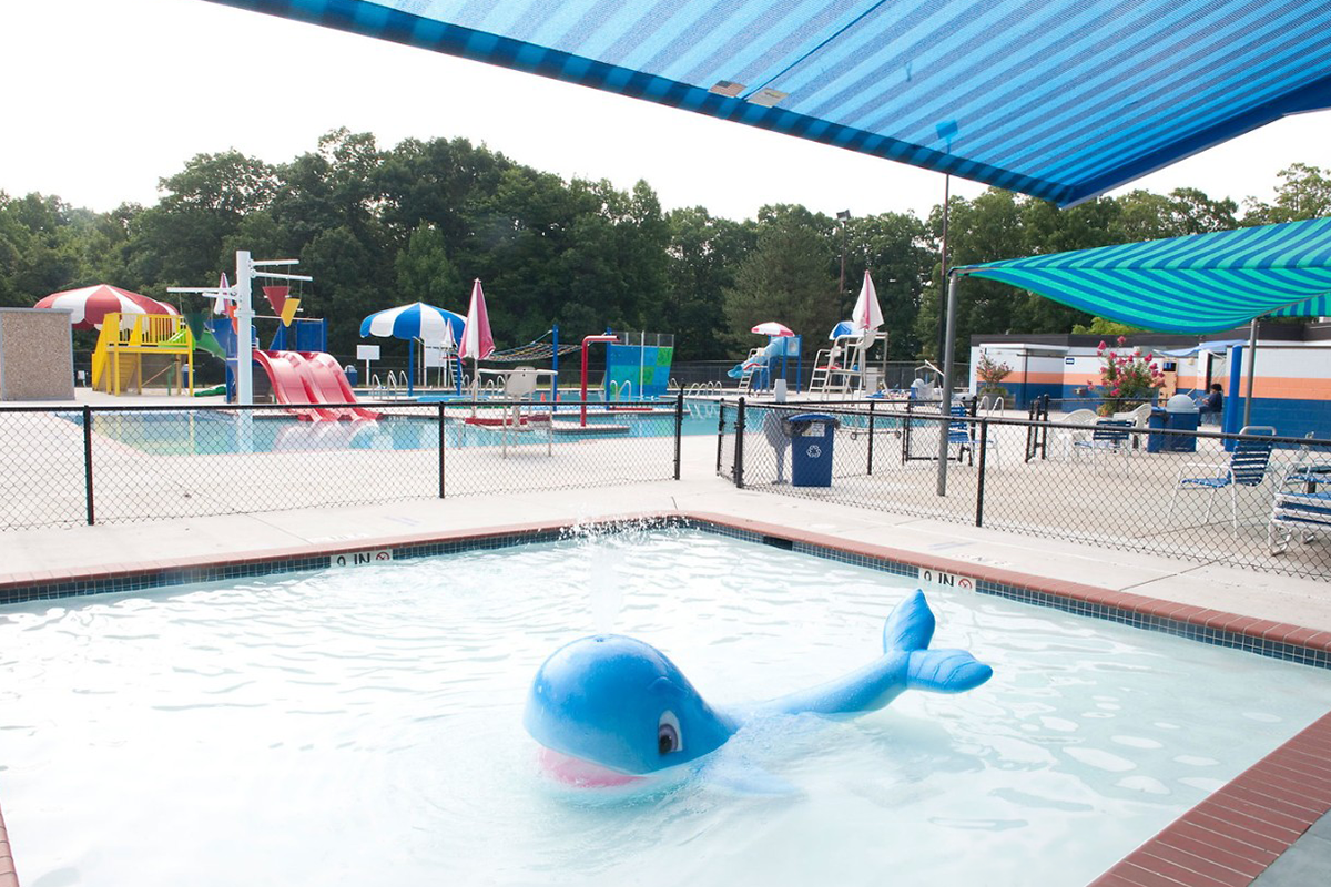 An outdoor kiddie pool with a striped blue awning above it and a blue whale-shaped fountain in the middle, spouting water.
