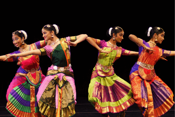 Four women wearing brightly colored dresses dancing on a dark reflective stage with a black background behind them