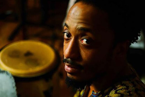 A man wearing a colorful, patterned tunic raises his eyebrows. Next to him is a traditional drum.