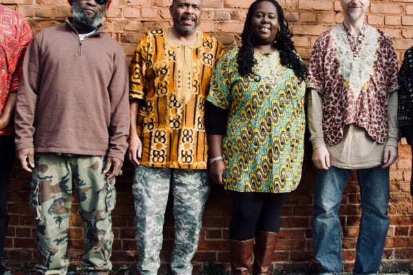 Four adults pose on a sidewalk with their backs against a brick wall. Most are wearing shirts made of colorful, patterned fabric.