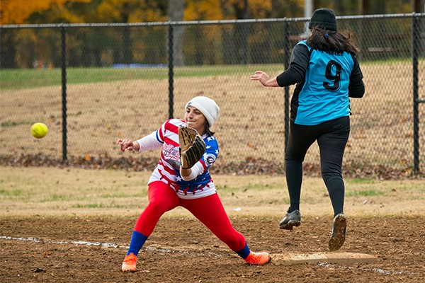 Two women playing softball. One is leaning down, bracing to catch a ball and tag first base. The other is running through first base. The batter/runner is safe