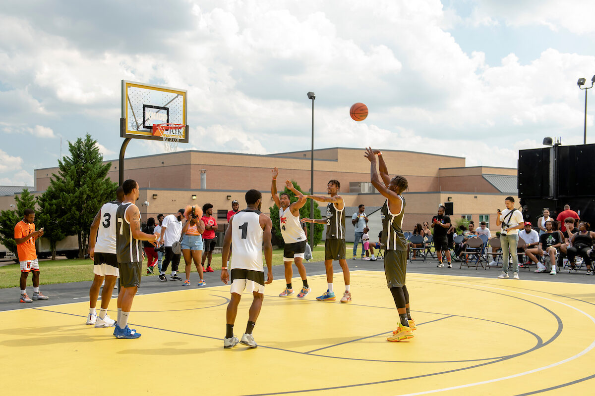 Men play basketball on an outdoor court with a bright yellow surface, while spectators watch from seats along the sidelines. In the background is a large brick building.