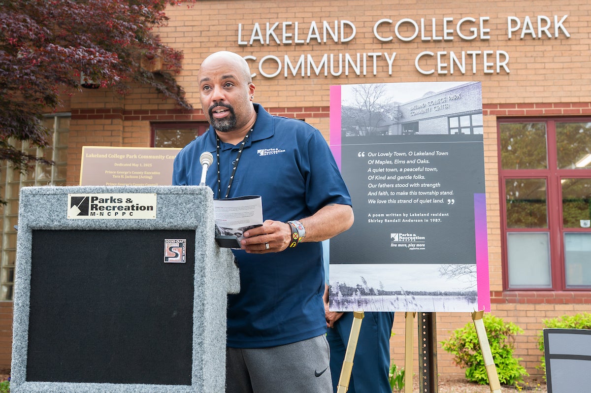 A man speaks at a gray podium outside in front of a brick building with letters that read LAKELAND COLLEGE PARK COMMUNITY CENTER. Behind the man is a poster on an easel featuring a poem and an older, black-and-white photo of the same building.