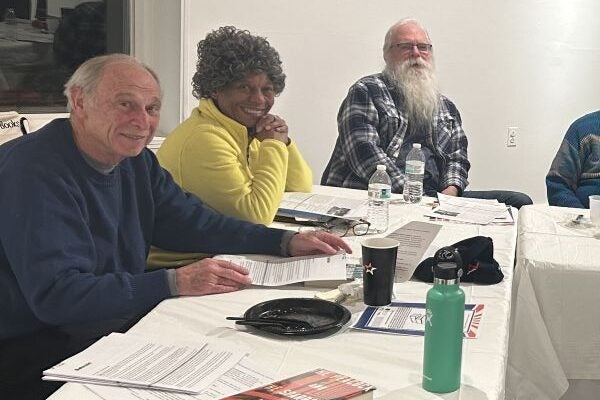 Three seniors sit at a long table covered in a white tablecloth. In front of them on the table are print-outs, books, and beverages.