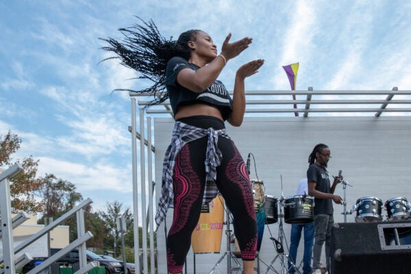 A woman with long hair dances on an outdoor stage. Behind her are multiple hand drums and a drum kit.