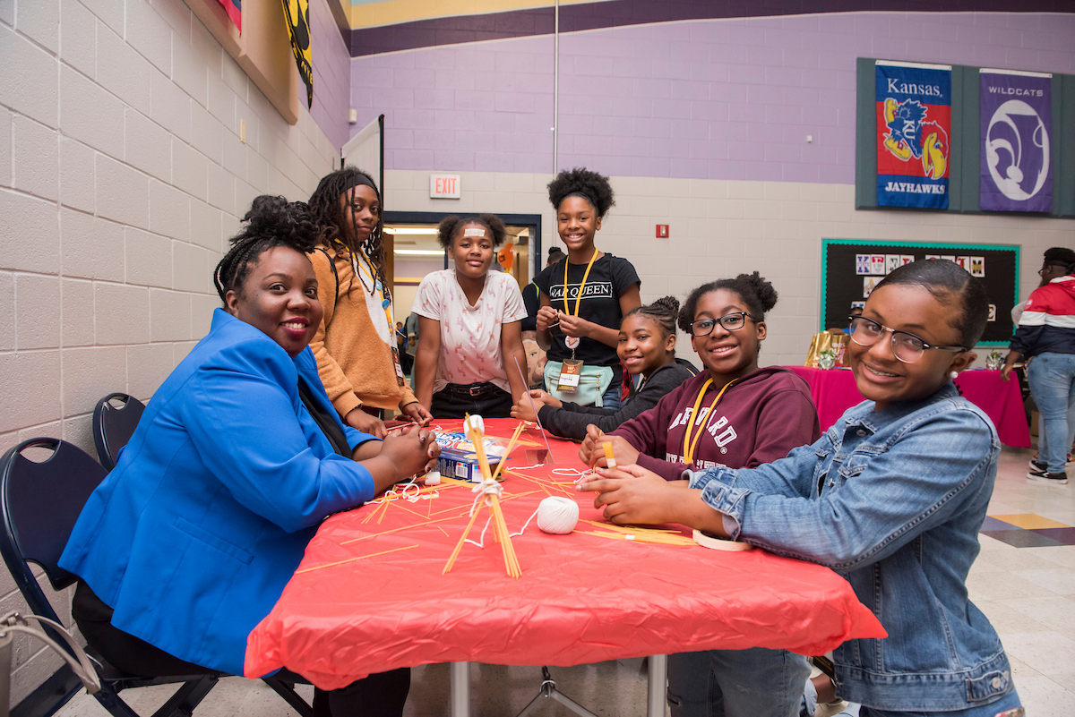 Inside a large event space with various college flags hanging on the walls, seven smiling young women sit around a long table making a craft project with uncooked spaghetti, string, and tape.