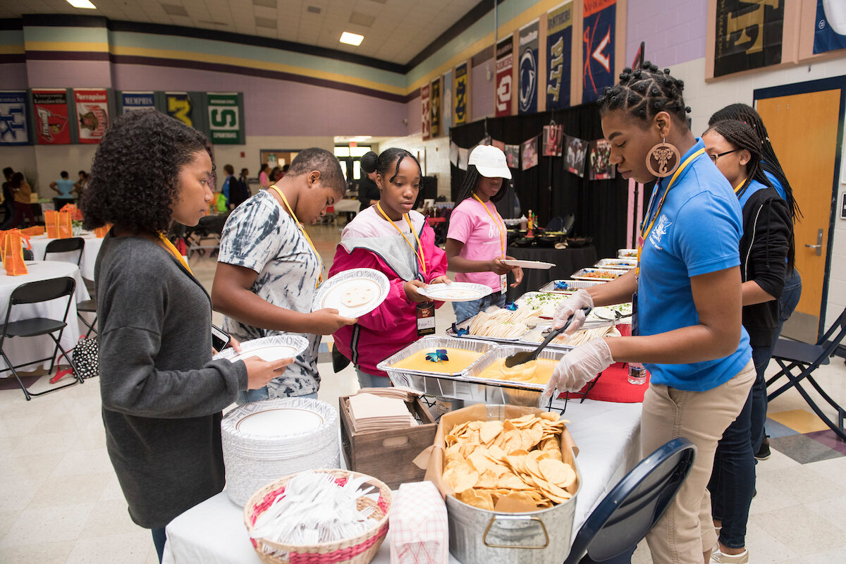 Inside a large event space with college flags hung around the walls, staff stand behind a table filled with buffet-style food, serving a line of young women holding paper plates. The room contains round banquet tables with orange gift bags at each place setting.