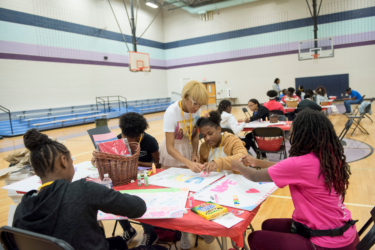 Inside a gymnasium, event tables are set up with various craft activities. At the nearest table, five young women use markers and paint to decorate large white posters.