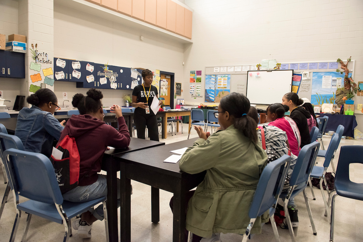 In a classroom, an instructor addresses a group of young women seated at tables.