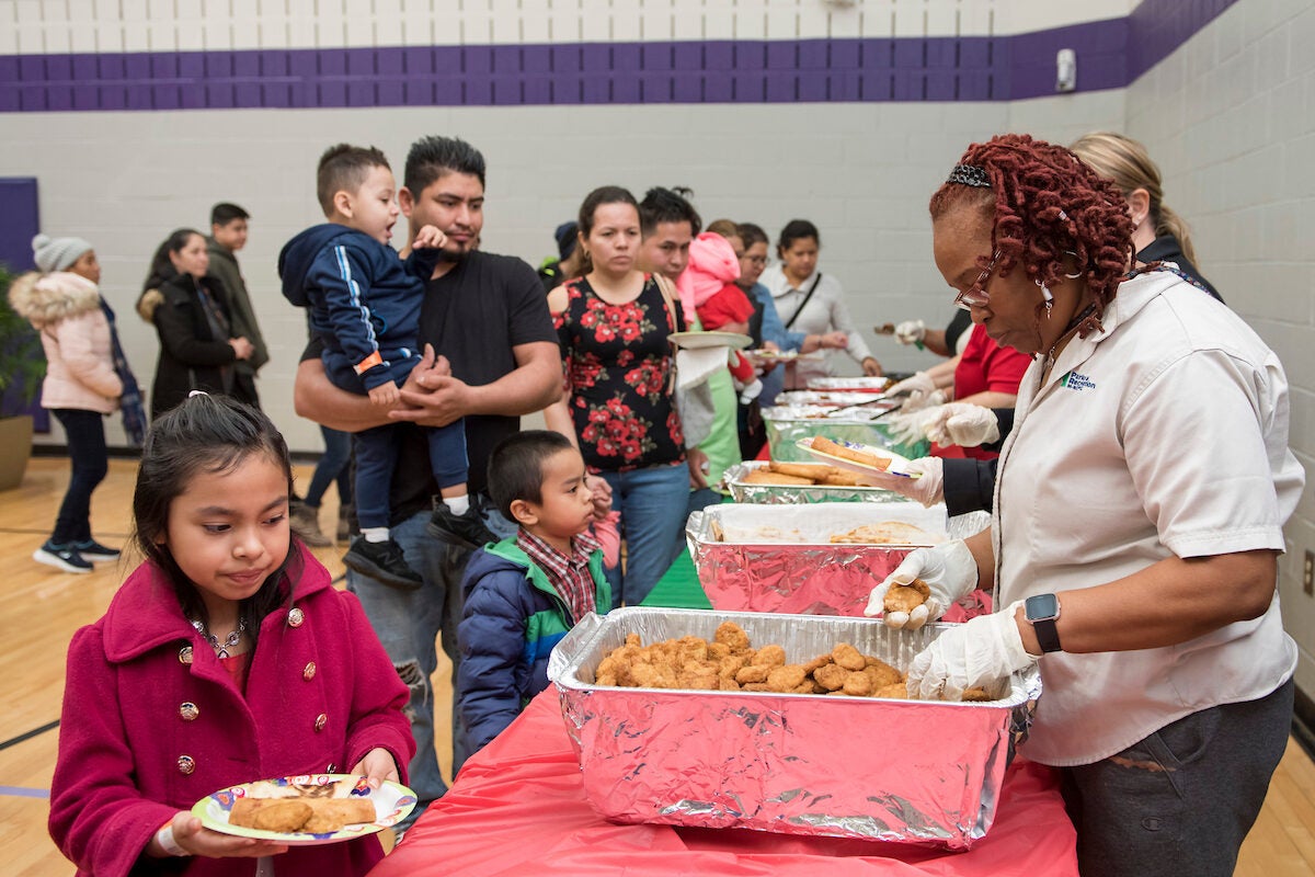 Inside a gymnasium with purple trim, adults in tan staff shirts stand behind long tables serving food from banquet dishes to a line of customers. The customers are mainly parents and small children, some wearing winter coats.