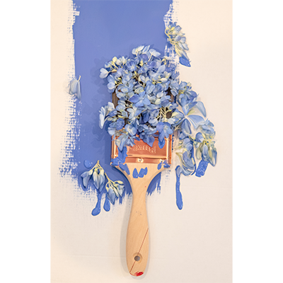 AS painting of a paintbrush creating a blue stroke on a surface. There are blue flower petals blossoming out of the paintbrush and paint on the surface