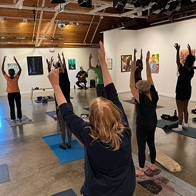 A group of people doing yogas on mats indoors. There is an instructor, as well as a man playing the guitar and playing jazz music.