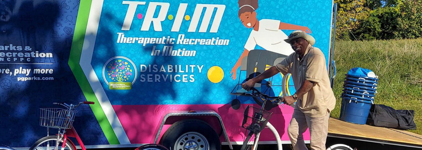 A smiling staff member in a tan Parks and Rec shirt stands on a white kickbike in a parking lot. Next to him is a large truck or trailer with the words TRIM: Therapeutic Recreation in Motion on the side, and the Parks and Rec logo. A red kickbike stands on the ground nearby.