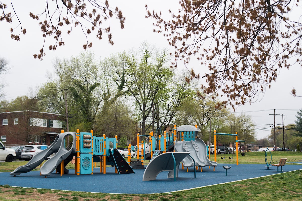 An outdoor playground with multiple play structures, featuring slides, platforms, stairs, climbing walls, musical instruments, games, spinning elements, and swings. Behind the playground are brick duplex houses.