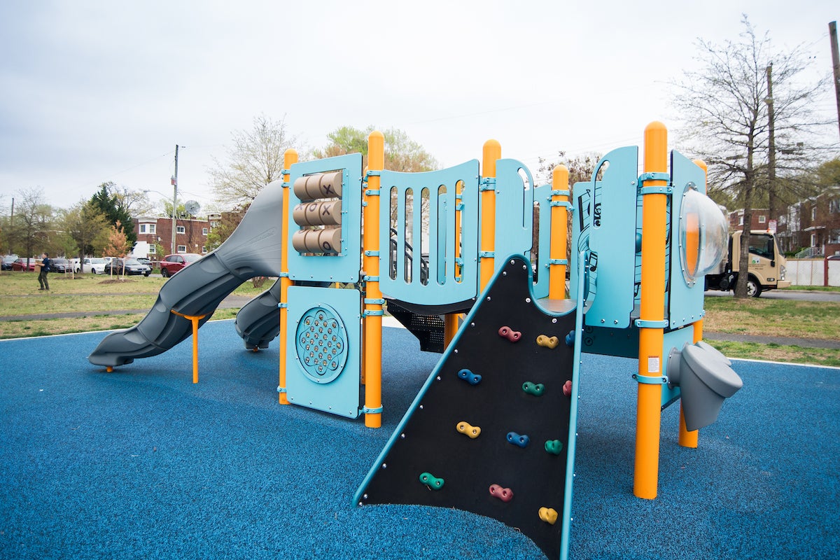 A large play structure at an outdoor playground, with slides, games, a climbing wall feature, multiple platforms and musical instruments. The play structure is light blue with yellow vertical posts.
