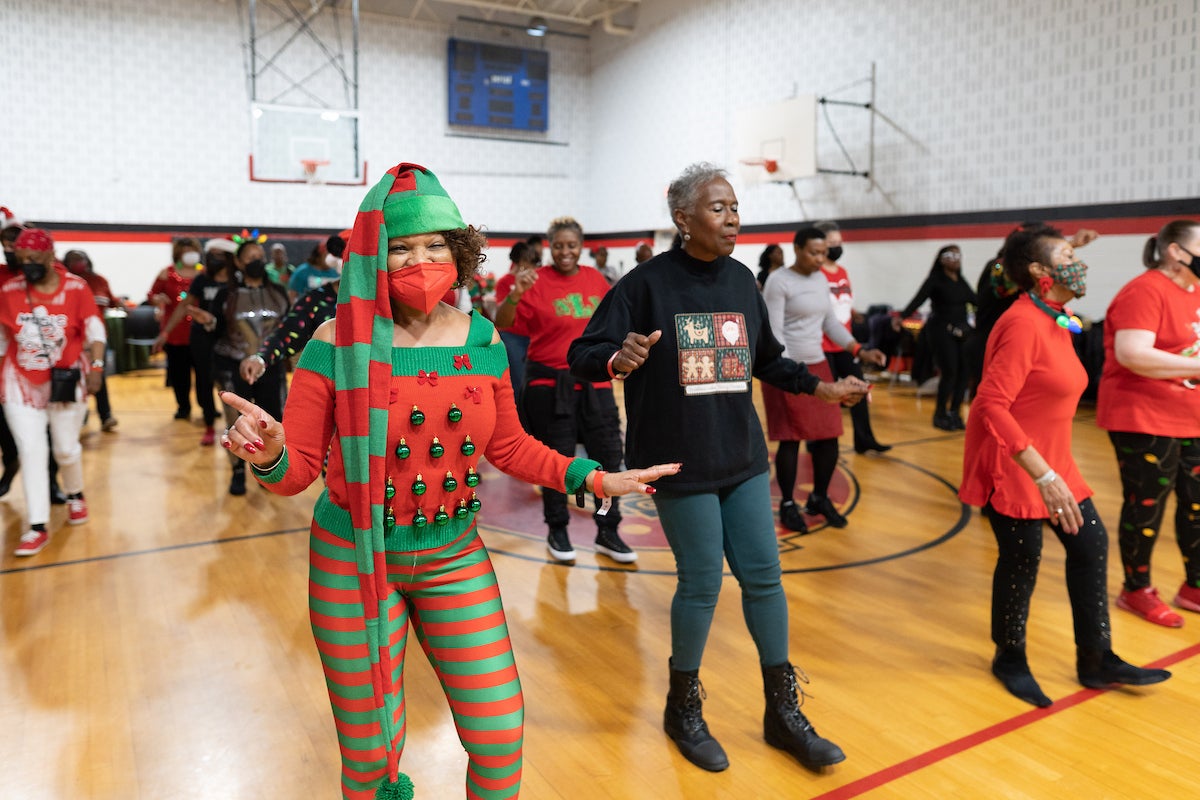 Several dozen adults in festive holiday clothing dance in formation inside a gymnasium. The woman closest to the camera is wearing red-and-green striped leggings and elf hat, and a red and green sweater decorated with holiday ornaments.