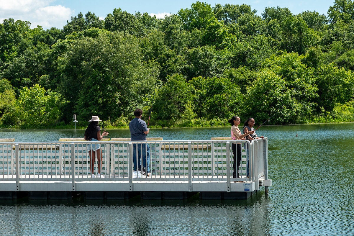 Four people fish with fishing poles off a white pier jutting out into a lake. The water is calm, with very tiny ripples from the breeze. Across the water is a forested area.