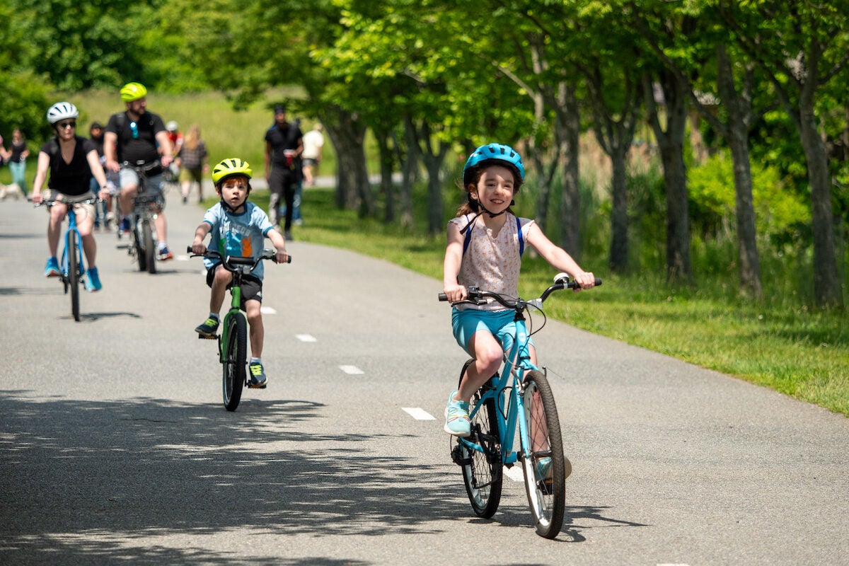 Two small children and their parents ride bikes on an asphalt trail with a dotted white line down the middle. There are people walking and jogging on the trail in the background, and someone walking a dog. The cyclist family all wear helmets that match the color of their bicycles.