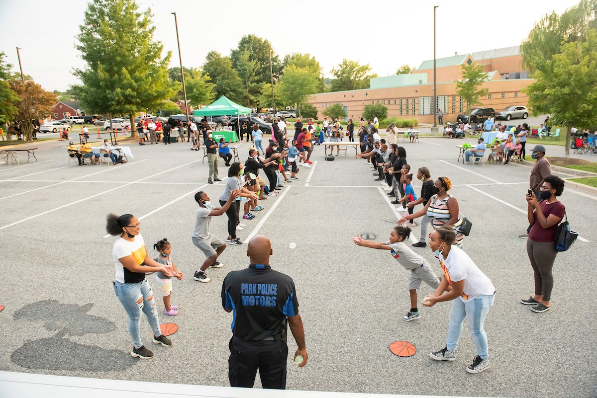 In a parking lot outside a large, brick building, two rows of people stand apart, tossing water balloons back and forth, at a community event.