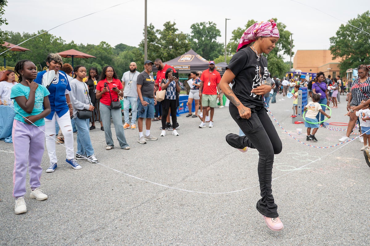 A woman wearing a pink scarf over her hair jumps inside two spinning ropes, double dutch style, in a parking lot, while dozens of people watch. Behind her are kids playing with hula hoops, lawn-sized Connect Four, and other games. There are event tents set up in a line leading back through the parking lot to a large brick building.